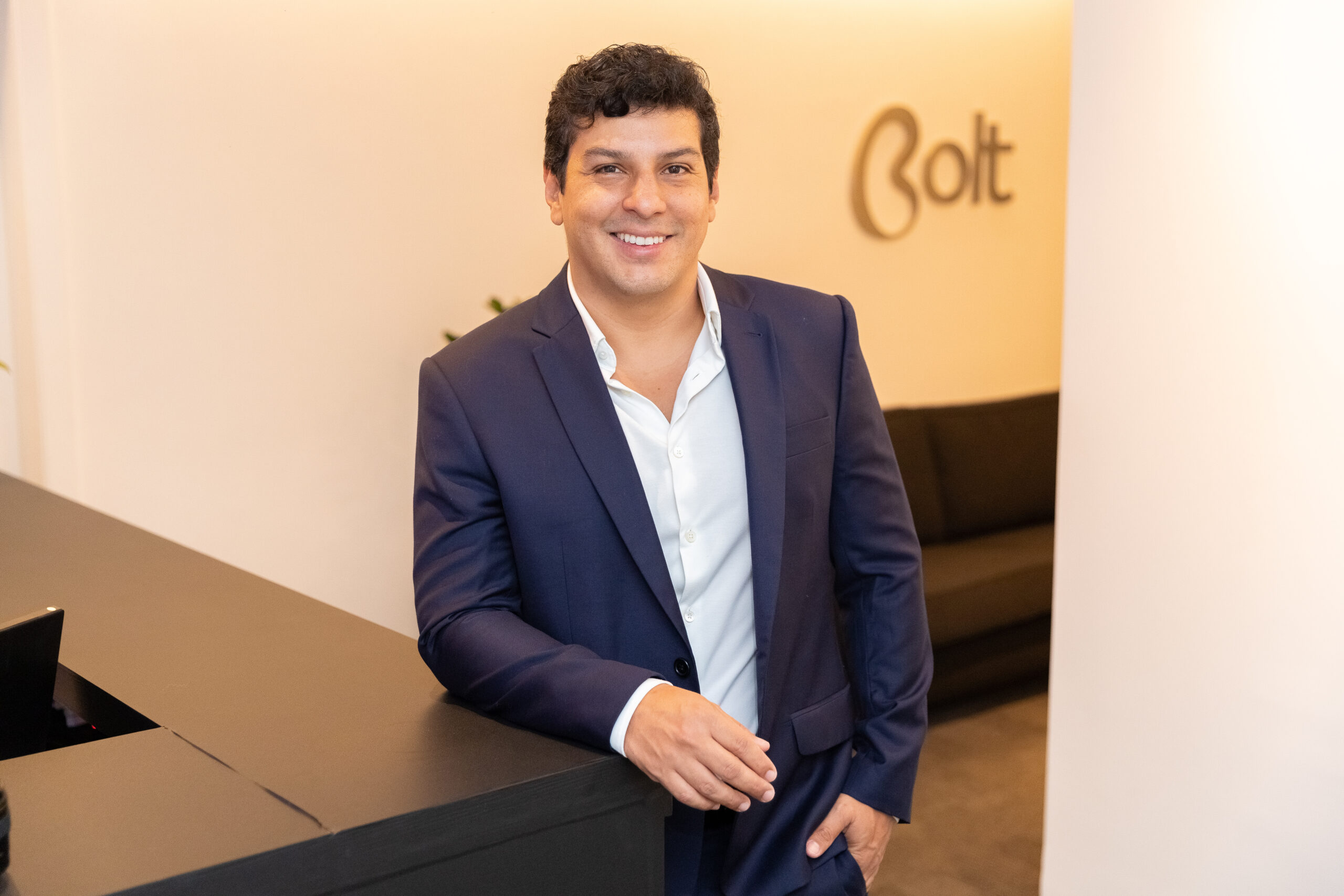 Temos a foto do Gustavo Ayala, CEO do Grupo Bolt. Ele é um homem de pele clara e cabelo escuro. Ele se apoia sob uma mesa preta e está usando camisa social e terno azul. Atrás dele, há um sofá preto e o letreiro Bolt na parede do escritório.