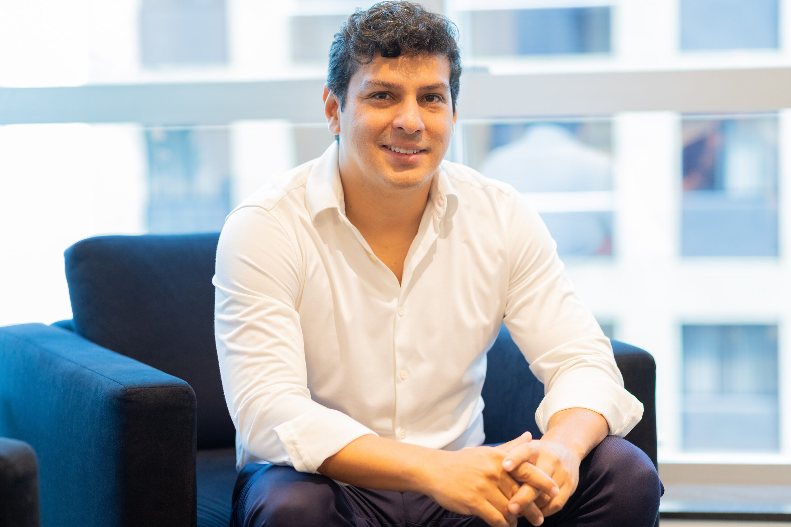 Temos a foto de Gustavo Ayala, CEO do Grupo Bolt. Ele é um homem de pele clara e cabelo escuro, curto. Usa camisa social branca e calça azul. Está sentado em um sofá azul e atrás dele, por meio de uma janela de vidro, vê-se prédios.